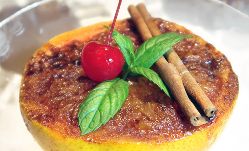 A plated grapefruit brûlée dessert topped with mint leaves, a cherry and a cinnamon stick.